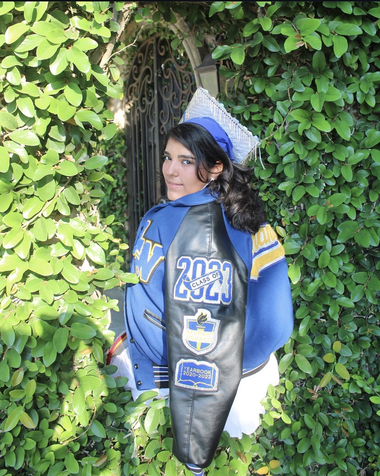 Graduate wearing a blue letterman jacket and decorated graduation cap among greenery
